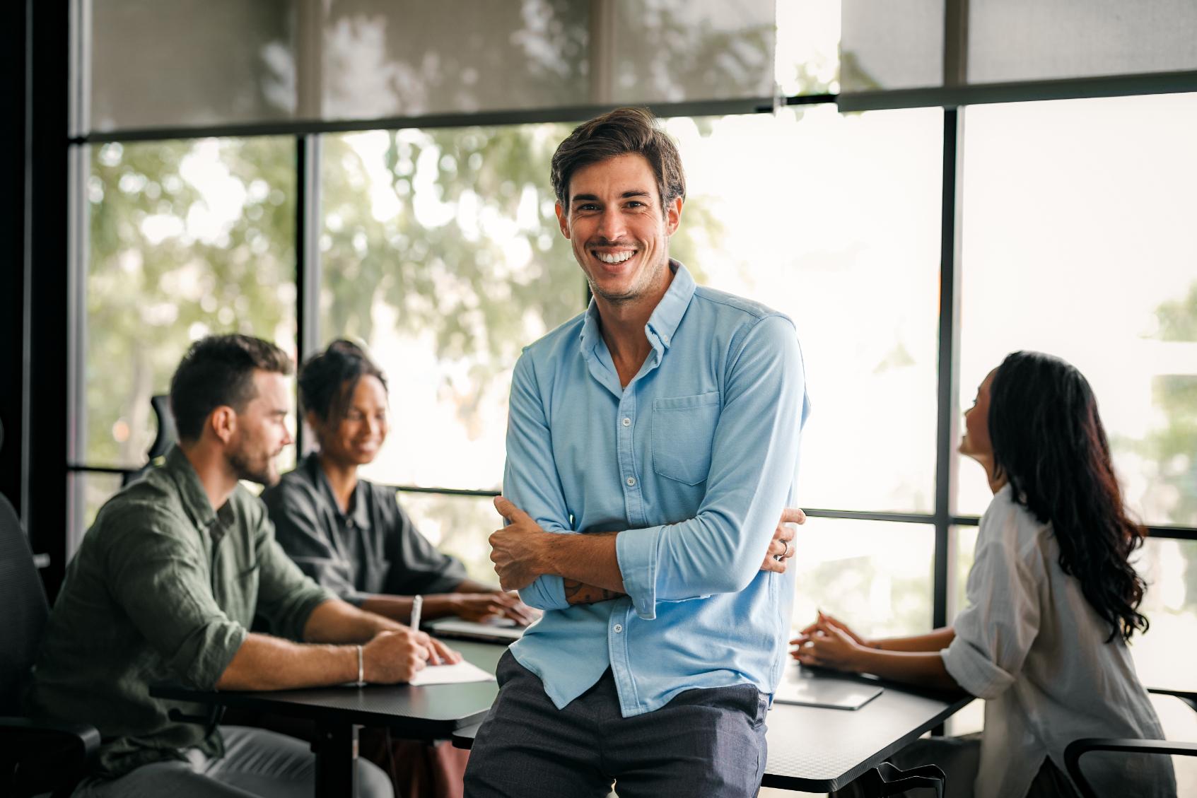 Smiling software engineer standing in a bright meeting room while his development team works at the table behind him